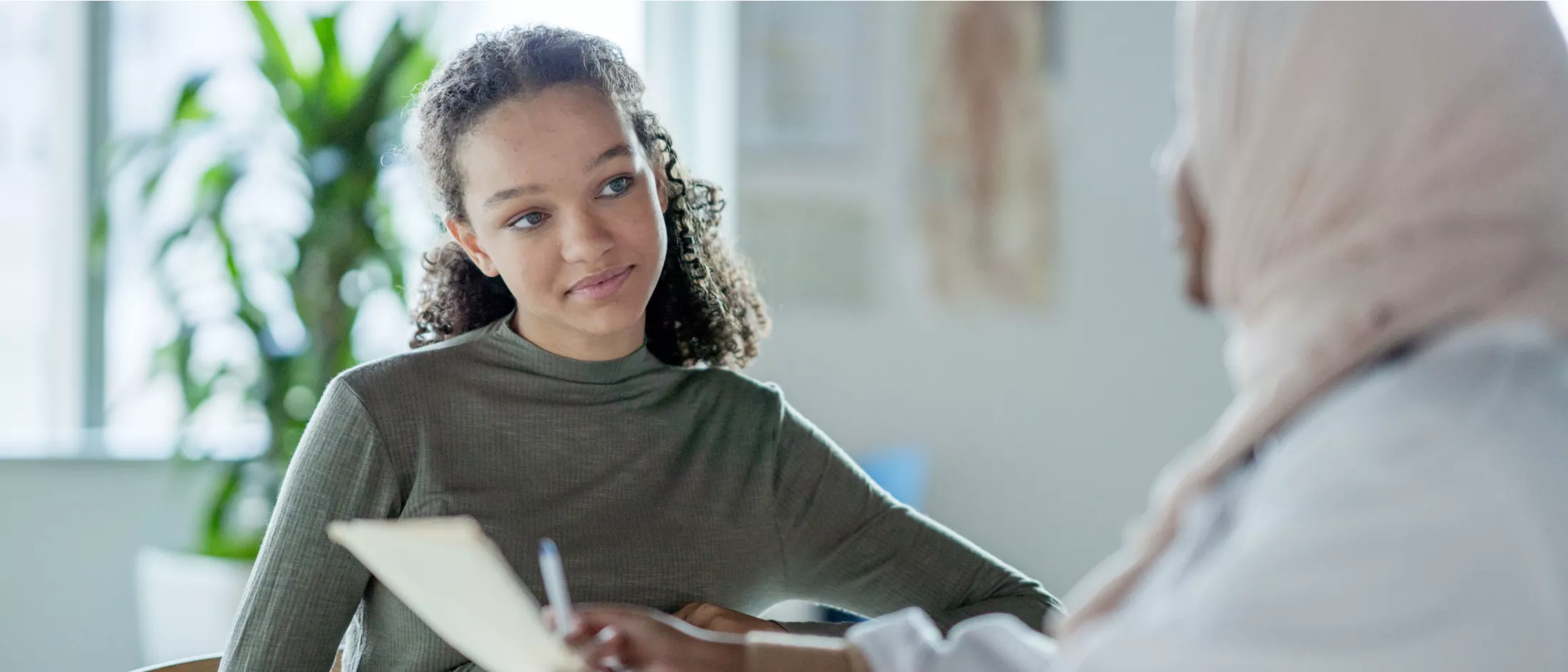 A black woman with long hair sits at a table and listens to a black woman wearing a headscarf and holding a document and pen.