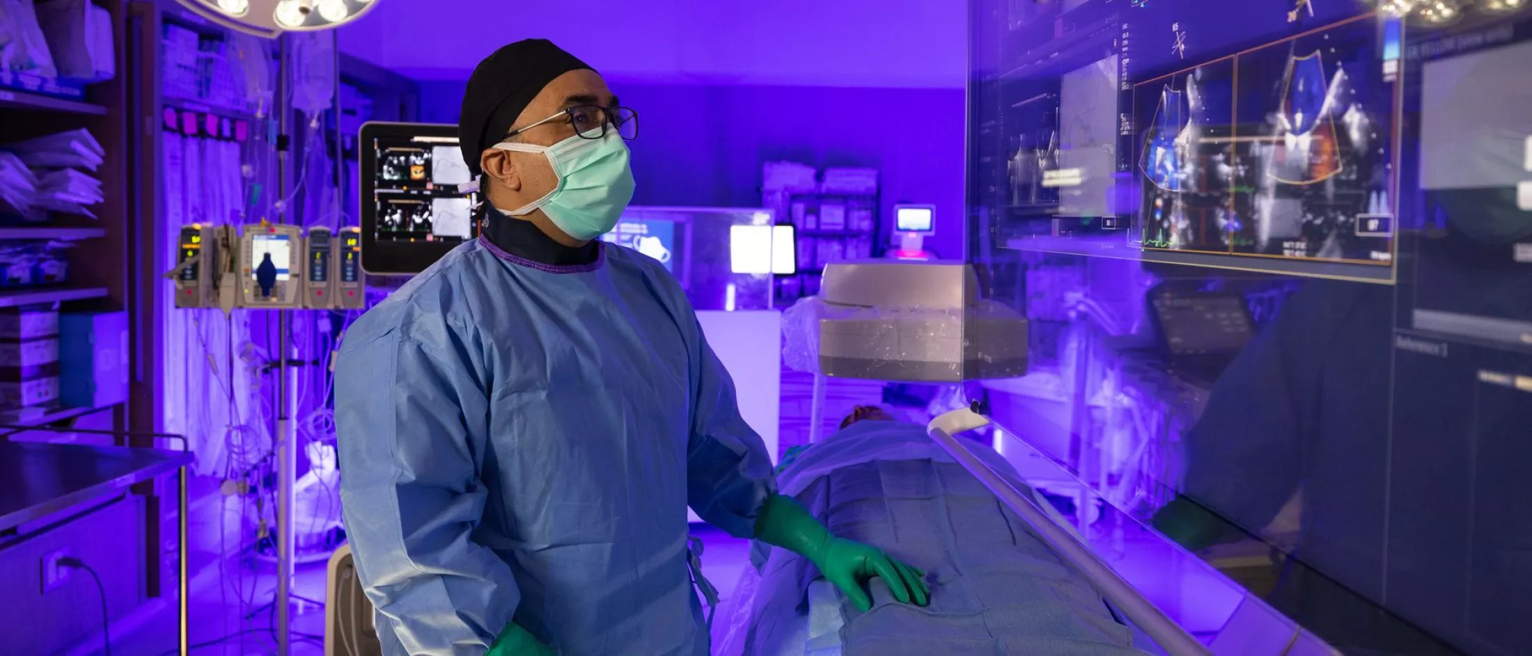 A doctor in a mask and full scrubs stands beside a patient on a table while looking at a monitor.