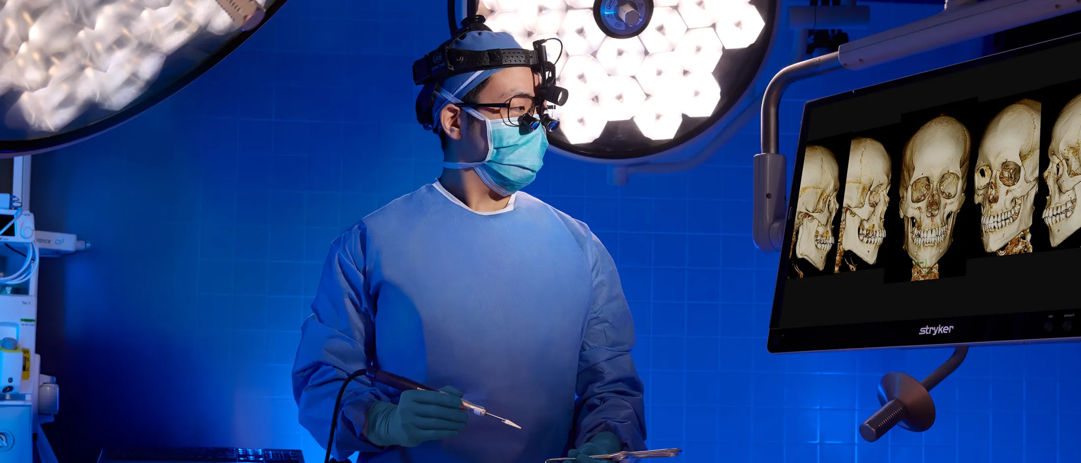 Surgeon wearing surgical loupes and mask, preparing instruments while viewing skull CT scans on a monitor.