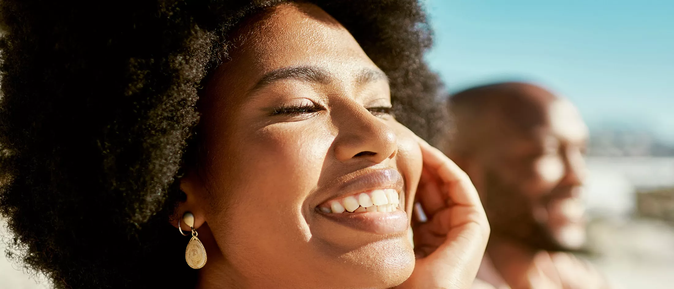 Woman smiling joyfully in the sunlight with a man blurred in the background.