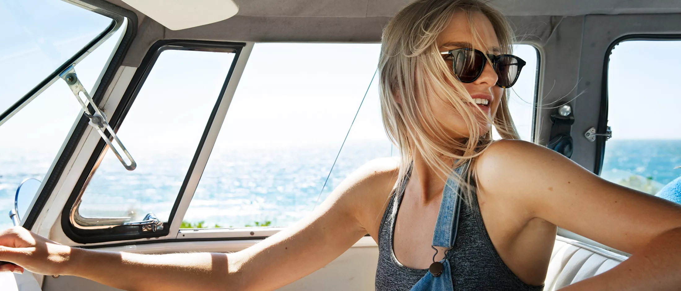 Woman wearing sunglasses and overalls driving a vintage car by the ocean.