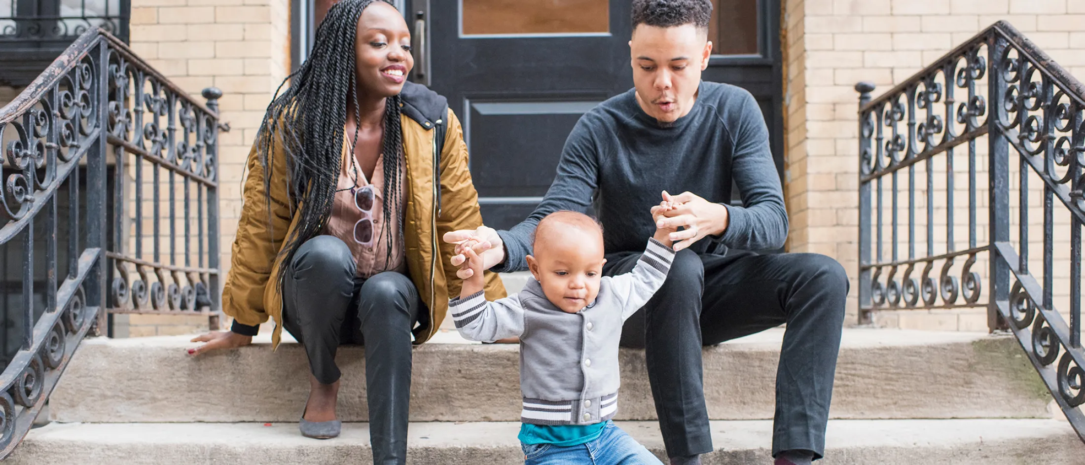 Young parents helping their toddler walk down steps outside their home, smiling and enjoying time together.