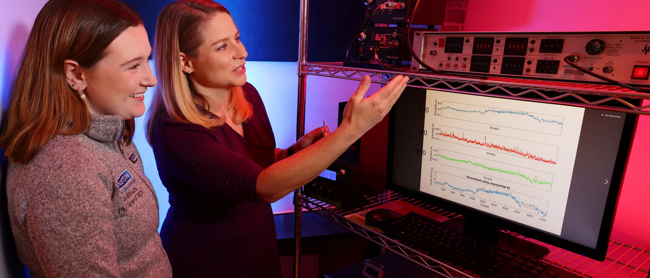 Two researchers analyze brainwave data on a computer screen surrounded by lab equipment and colored lighting.