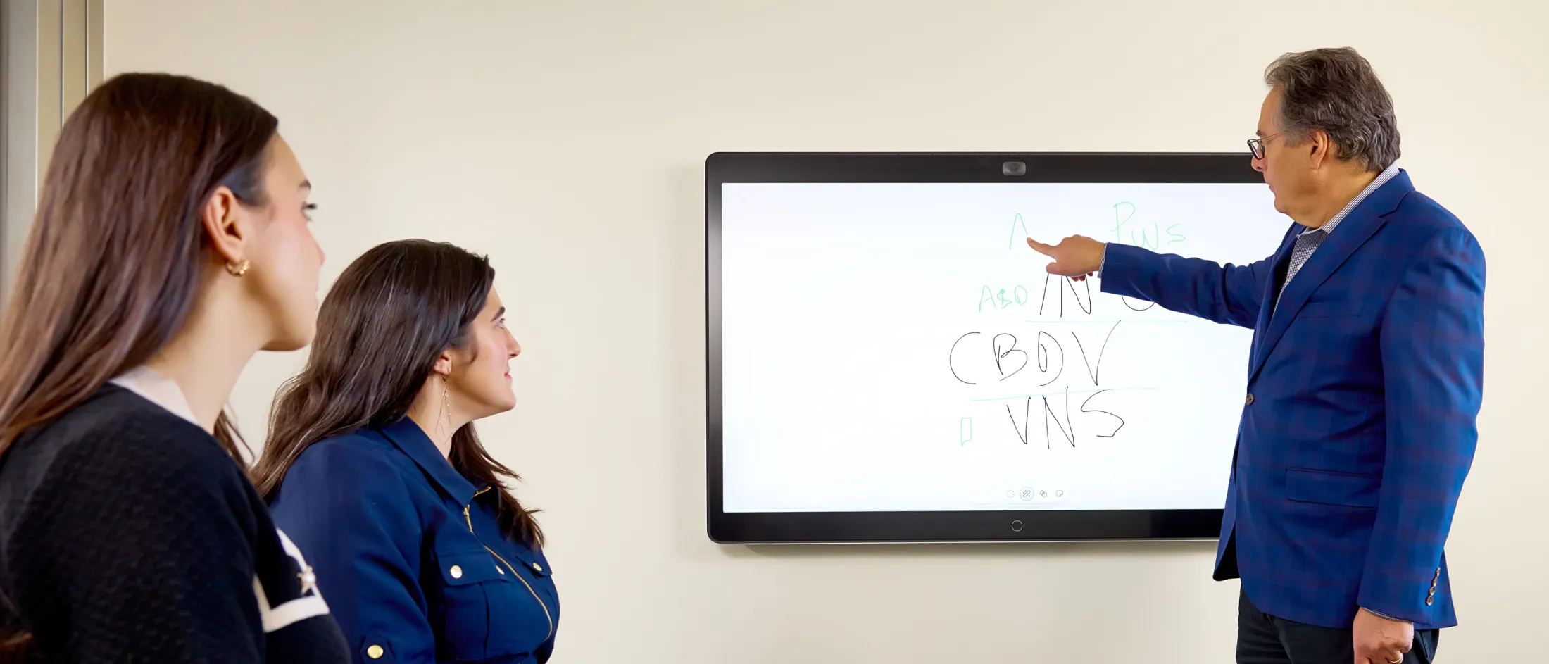Man in blue blazer points at a digital whiteboard while two women watch.