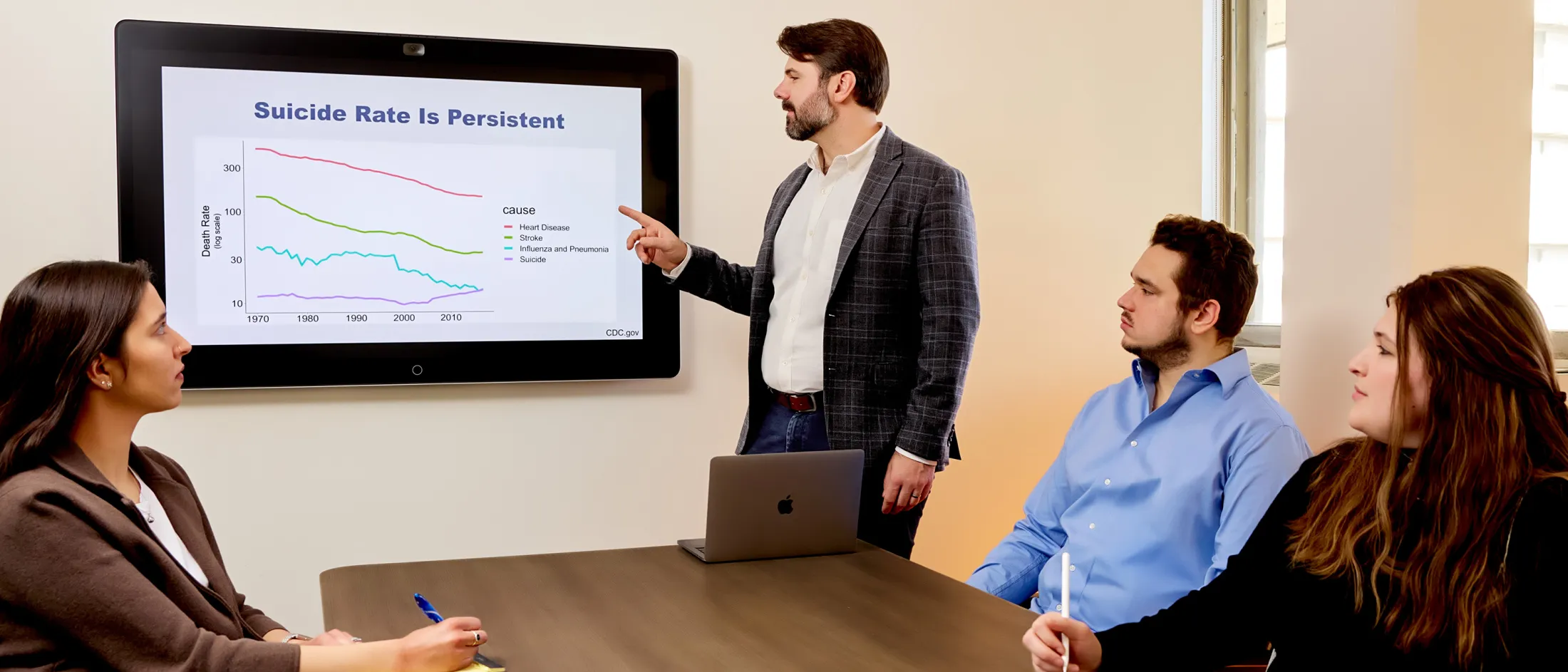 Man points to screen with suicide rate chart while three people listen during a meeting.