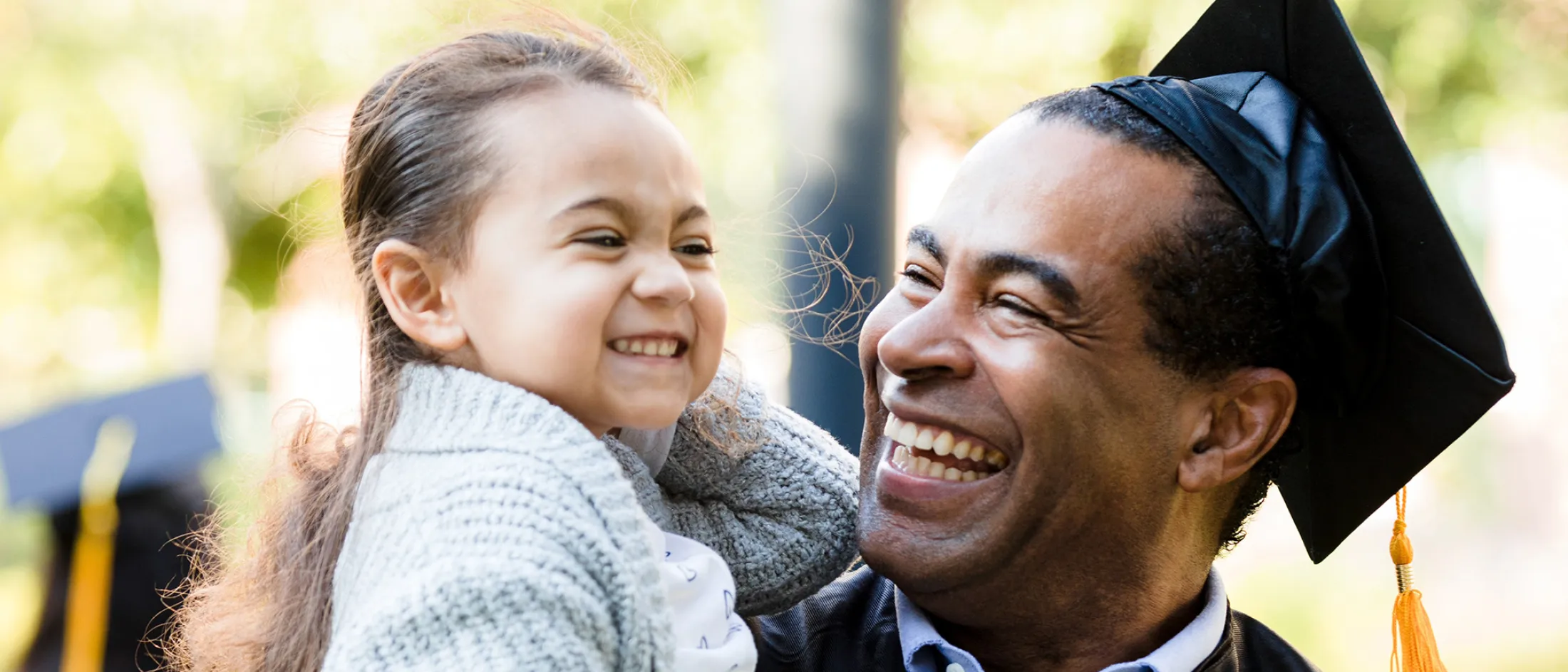 Father in graduation cap and gown holding his smiling daughter during a celebration outdoors