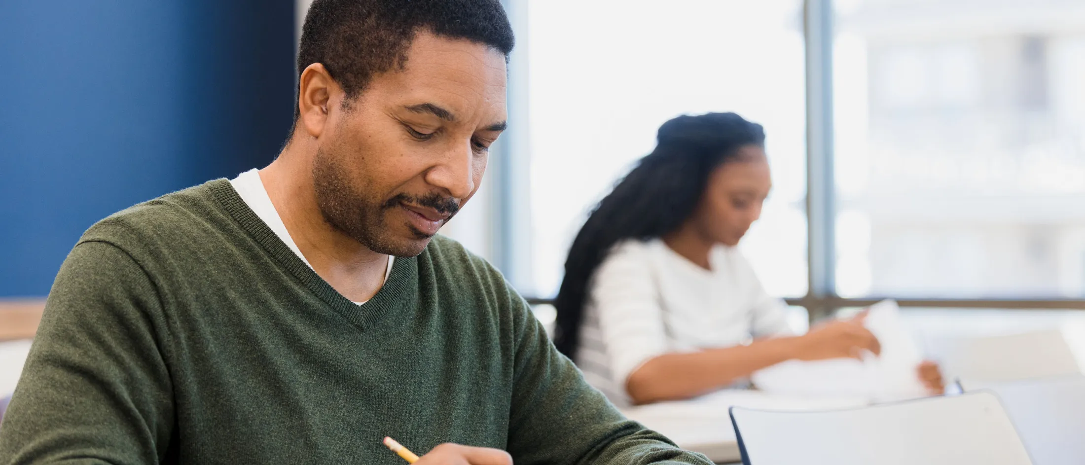 Man writing on paper in a classroom while a woman studies in the background