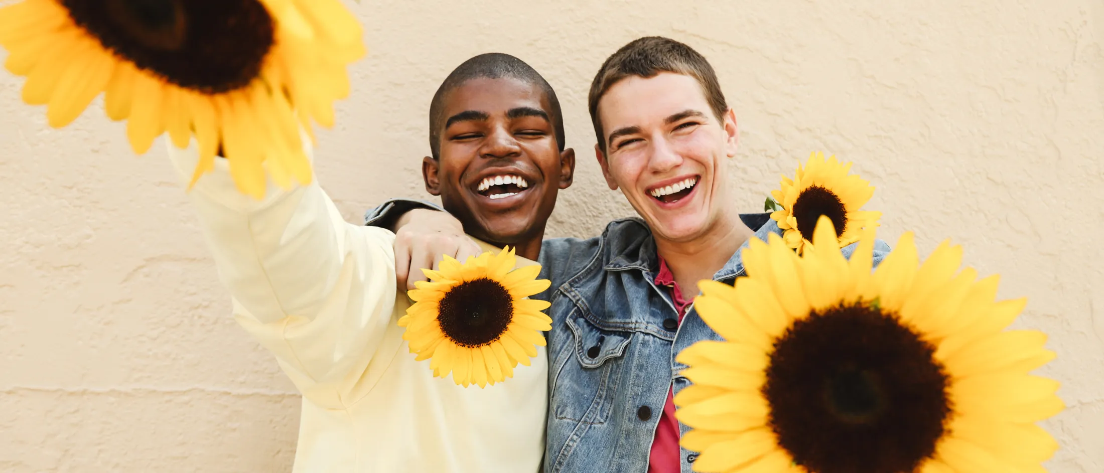 Two friends smiling and holding sunflowers, celebrating together in front of a beige wall
