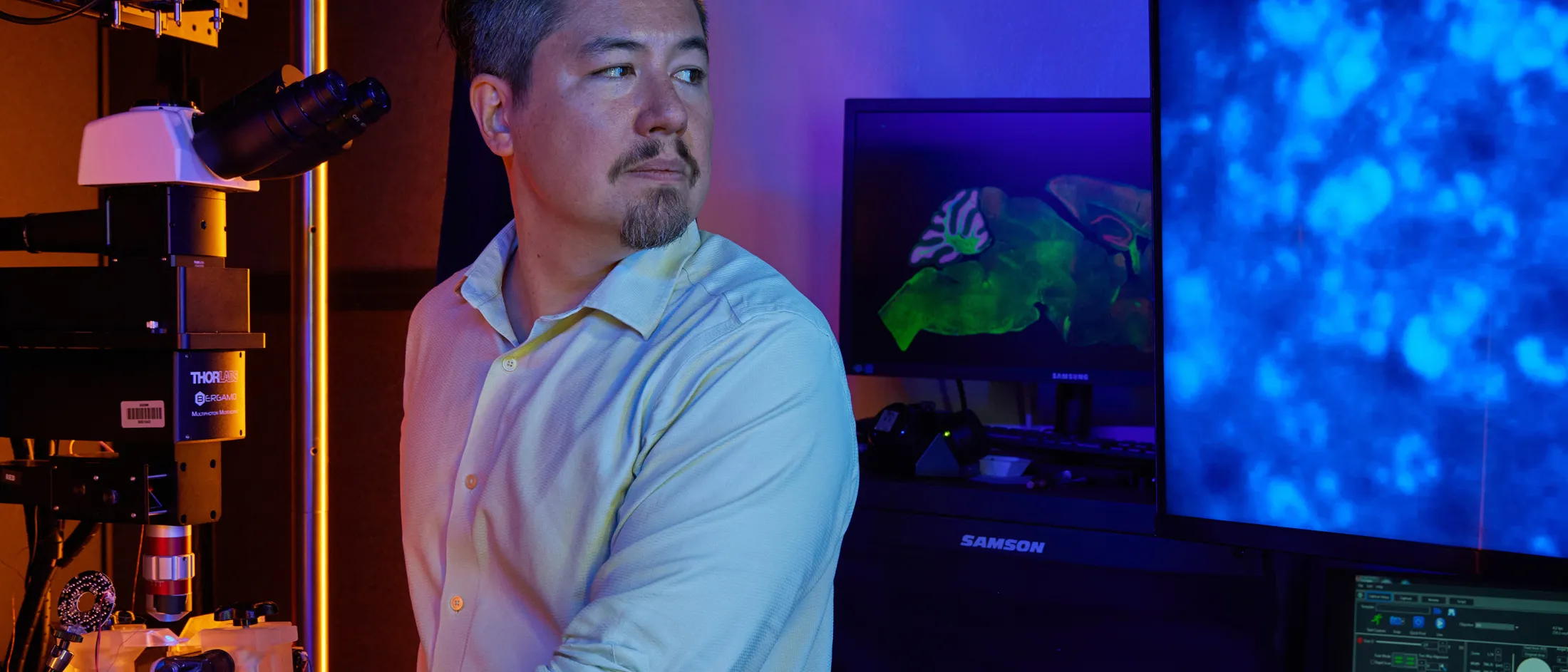 Researcher in lab beside microscope and glowing screens displaying brain scan visuals.