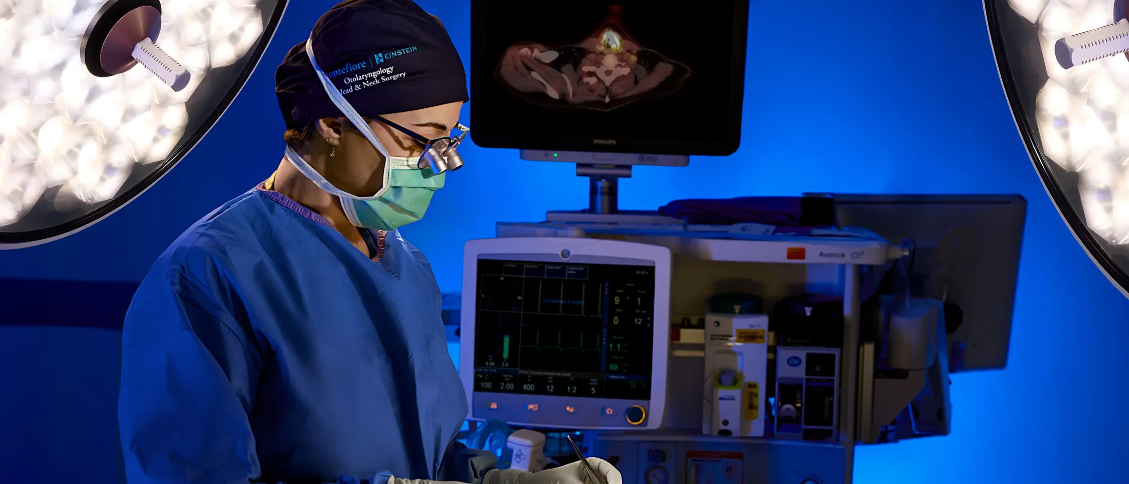 A masked surgeon in a blue operating room, focused on a procedure with medical monitors and lights illuminating the scene.