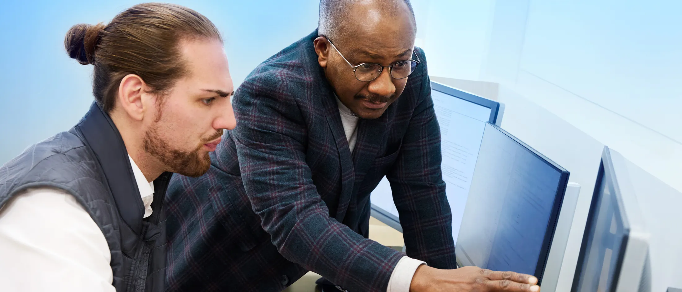 Two men collaborate at a computer workstation, with one pointing at the screen while explaining something to the other.
