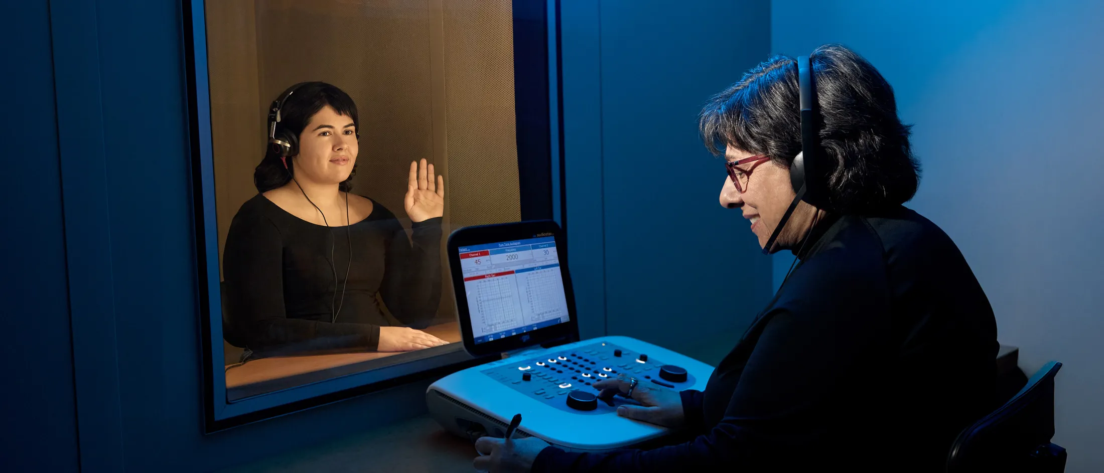 Audiologist conducting a hearing test with a patient seated behind glass, both wearing headsets.