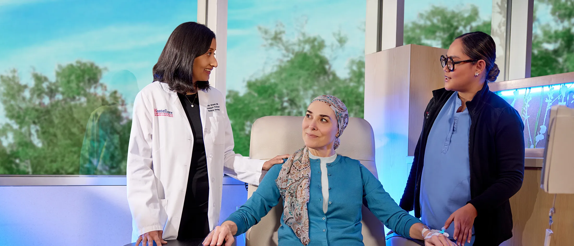 Female doctor and nurse standing beside a woman in a headscarf receiving treatment, smiling and offering support in a bright medical setting.