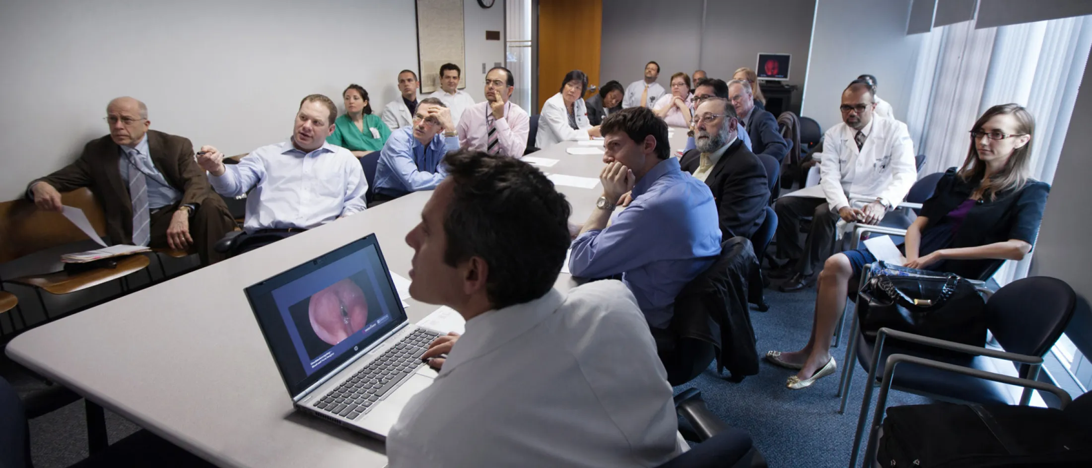 Group of medical professionals seated around a conference table viewing a medical image on a laptop during a tumor board meeting.