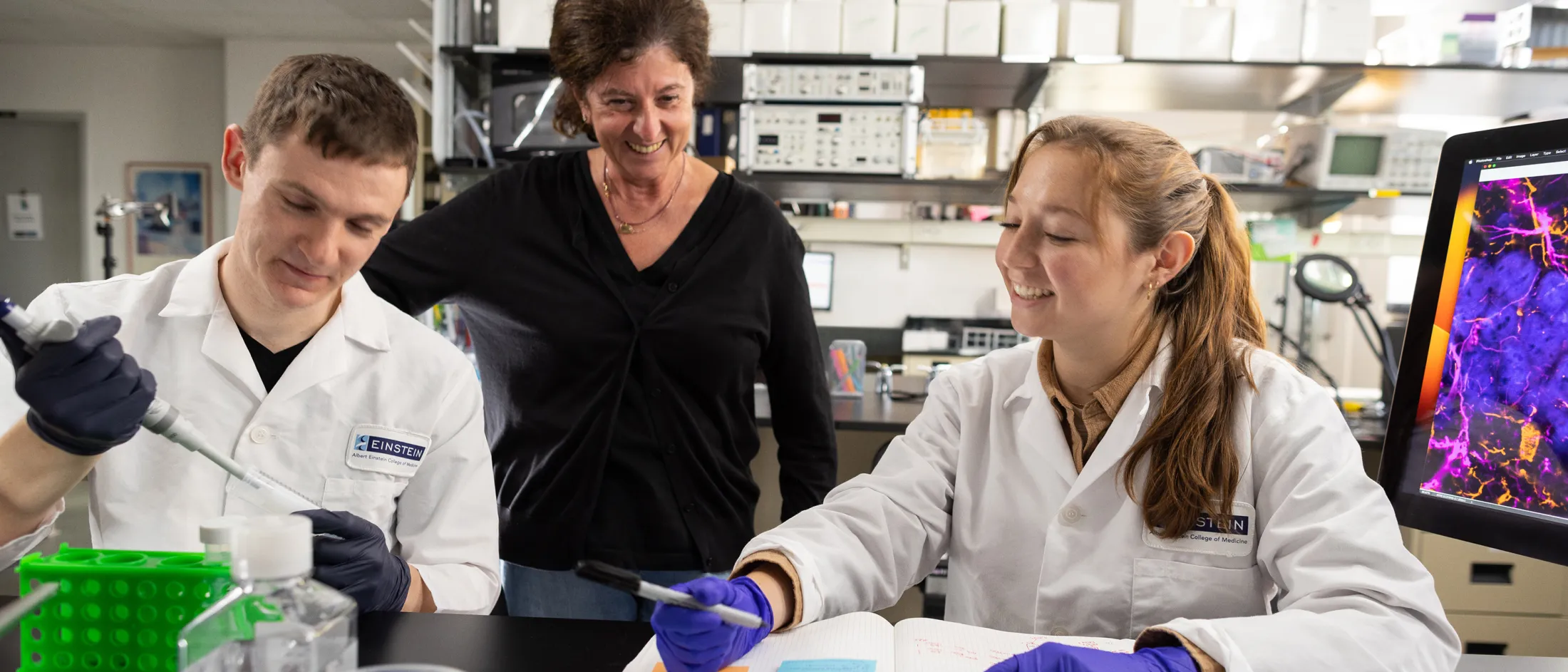 Two students in white lab coats working with lab equipment while a professor observes and smiles.