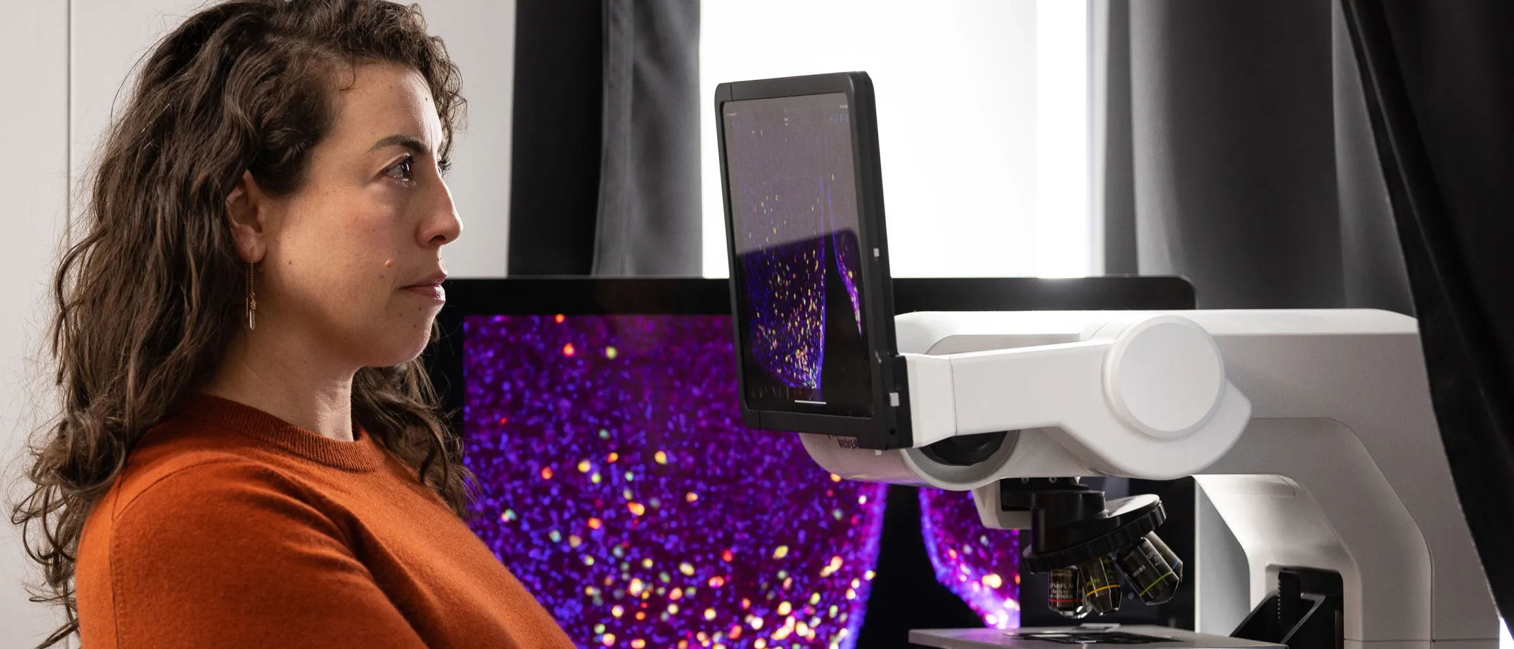 Scientist in orange sweater examining microscope images on a digital display in a research lab.