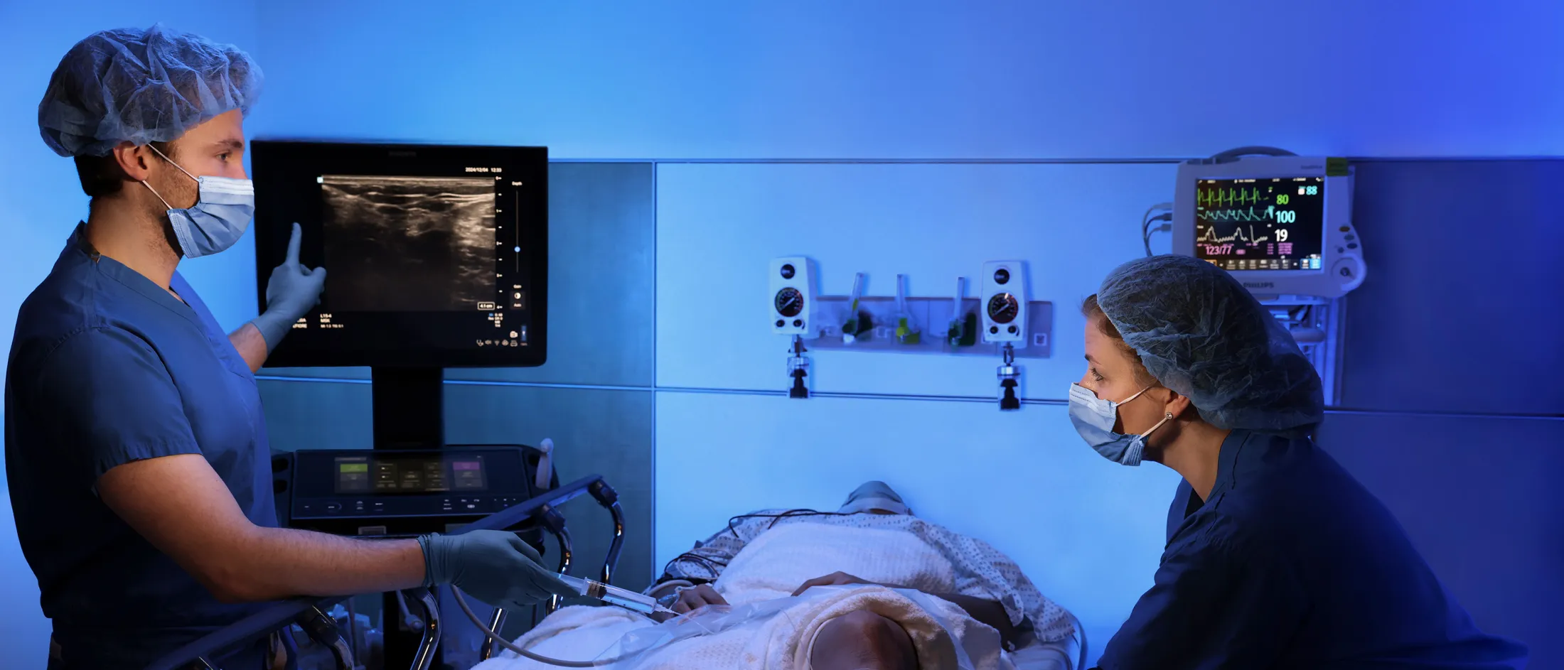 Medical staff using ultrasound equipment on a patient's leg in a modern hospital room with monitors and machines.