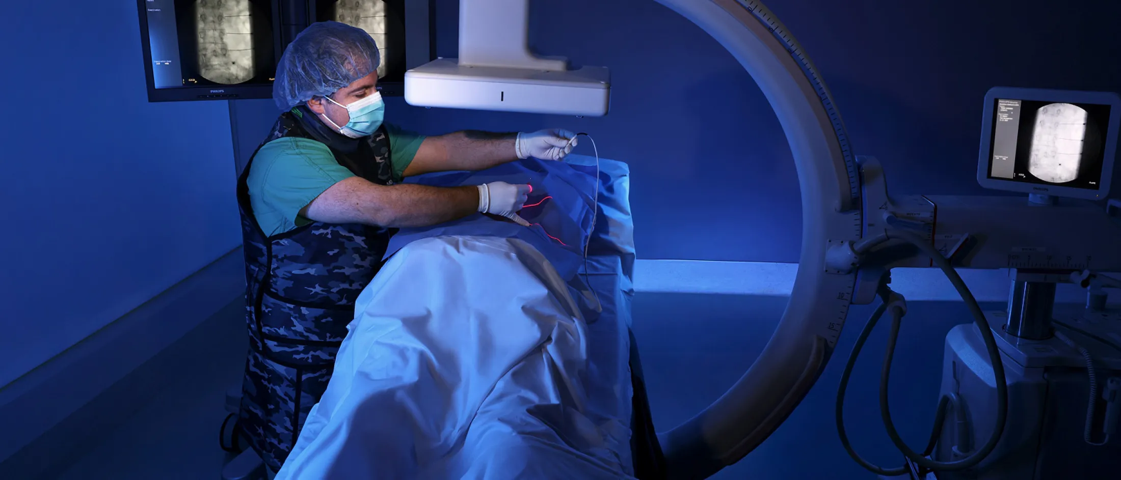 Doctor in surgical gear performing a procedure on a patient under an X-ray machine in a dimly lit medical room.