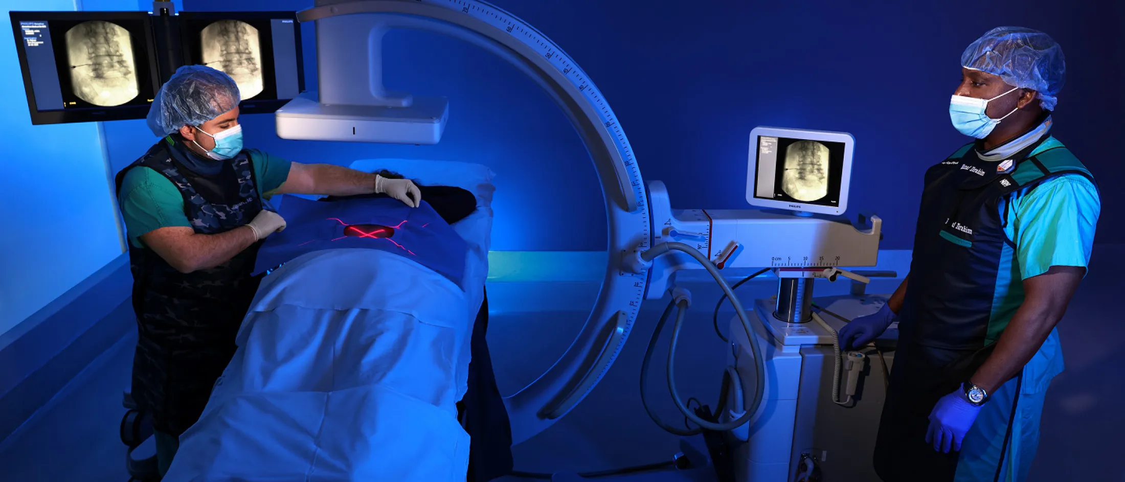 Two medical professionals conducting a procedure using a C-arm imaging machine, with one standing by the monitor and the other attending to the patient.