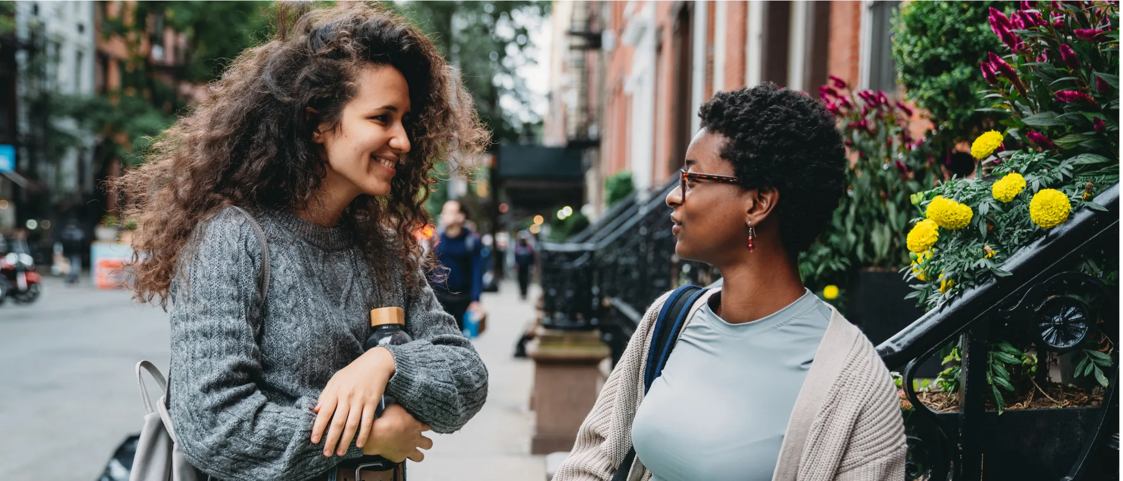 Two young women have a friendly conversation outside on a city sidewalk near a flower-filled stoop.
