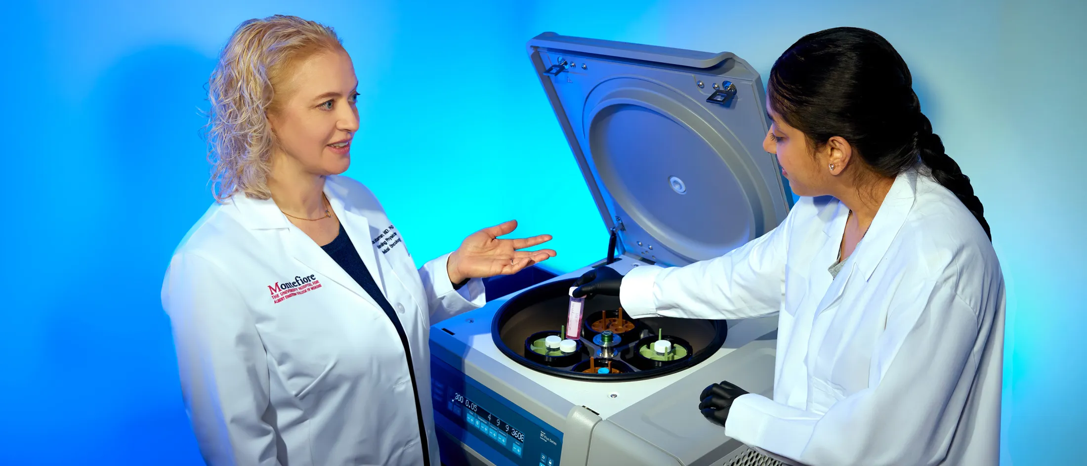Two female scientists in lab coats working with a centrifuge in a brightly lit lab.