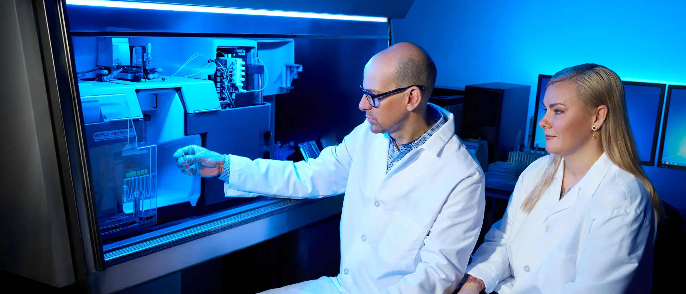 Male and female researchers in lab coats examining samples inside high-tech lab equipment under blue lighting.