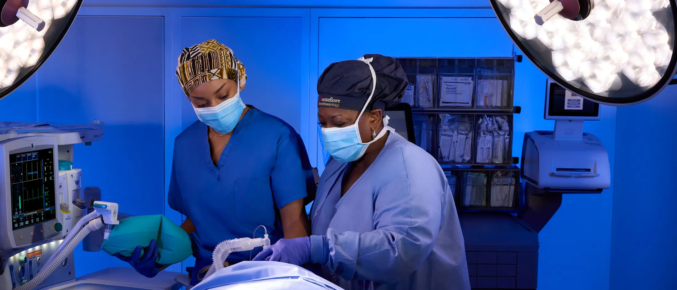 Two masked anesthesia providers preparing airway equipment beside an anesthesia machine in a blue-lit OR.