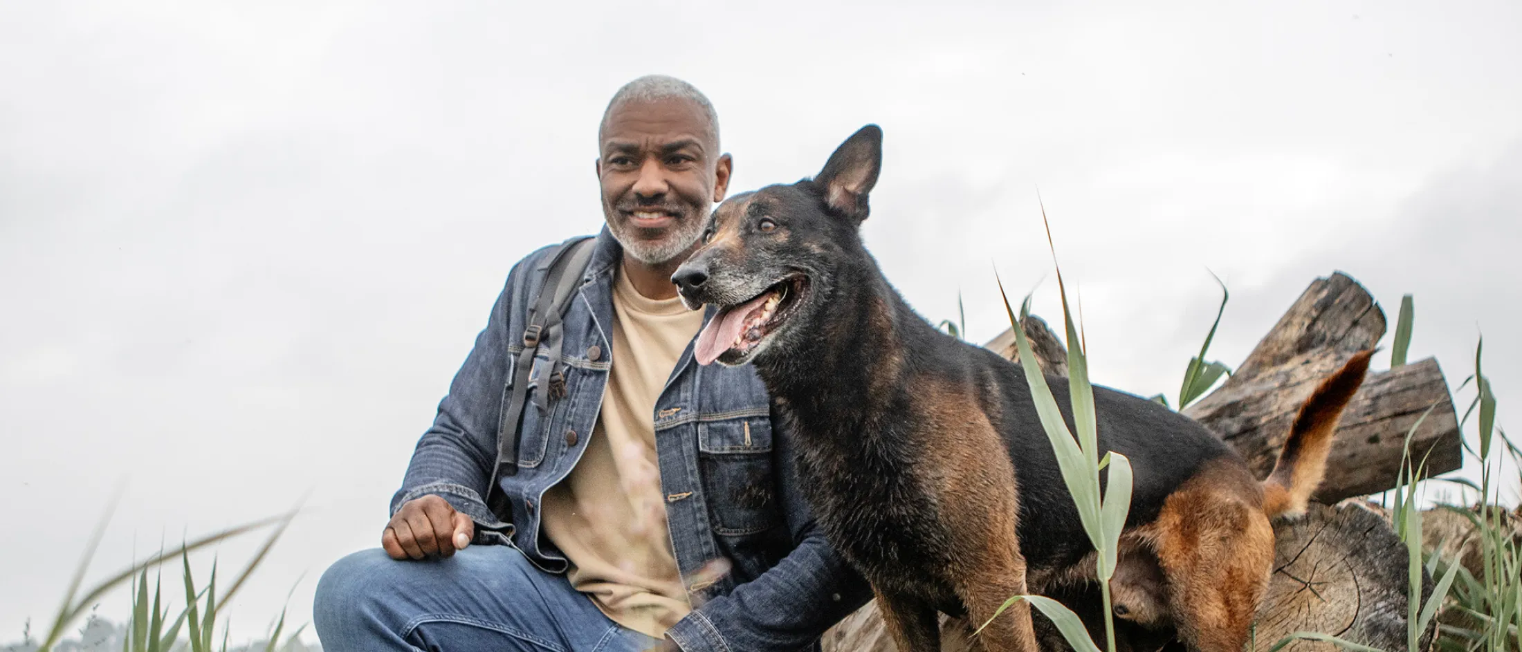 Man in denim jacket kneeling beside his happy large dog amid logs and tall grass outdoors.
