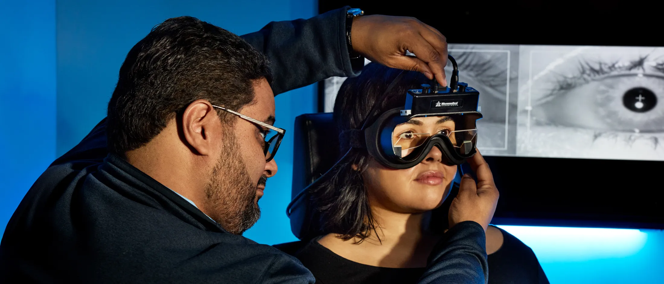 A doctor adjusts specialized diagnostic goggles on a seated female patient during a vestibular test, with an eye tracking screen in the background.