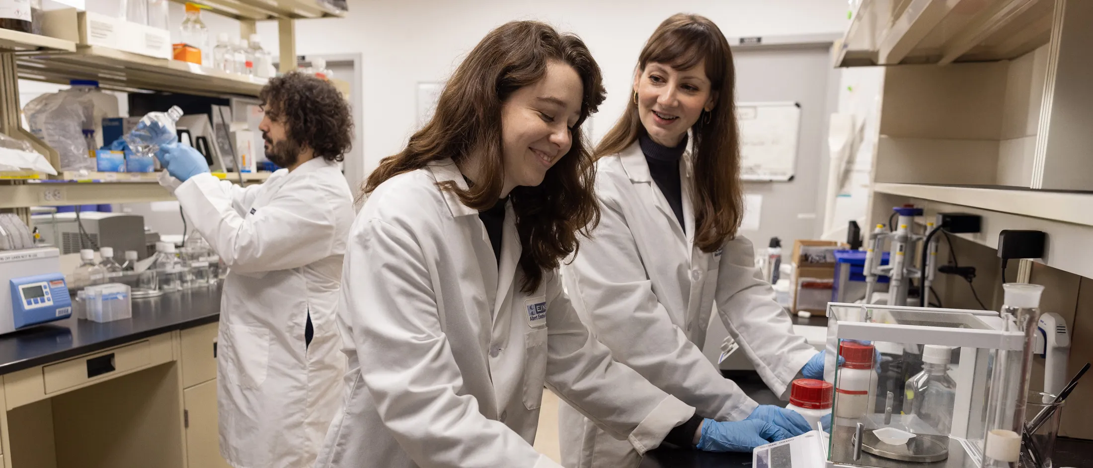 Three researchers in lab coats working in a laboratory; two women smiling while handling containers near a digital scale and a man examining a sample in the background.