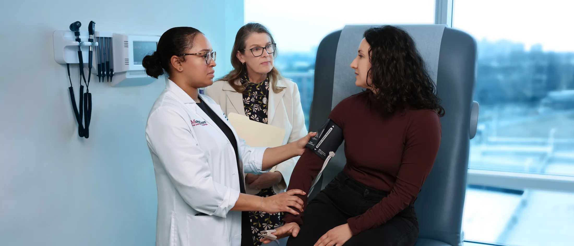 Doctor measuring a patient’s blood pressure in an exam room while another clinician observes.