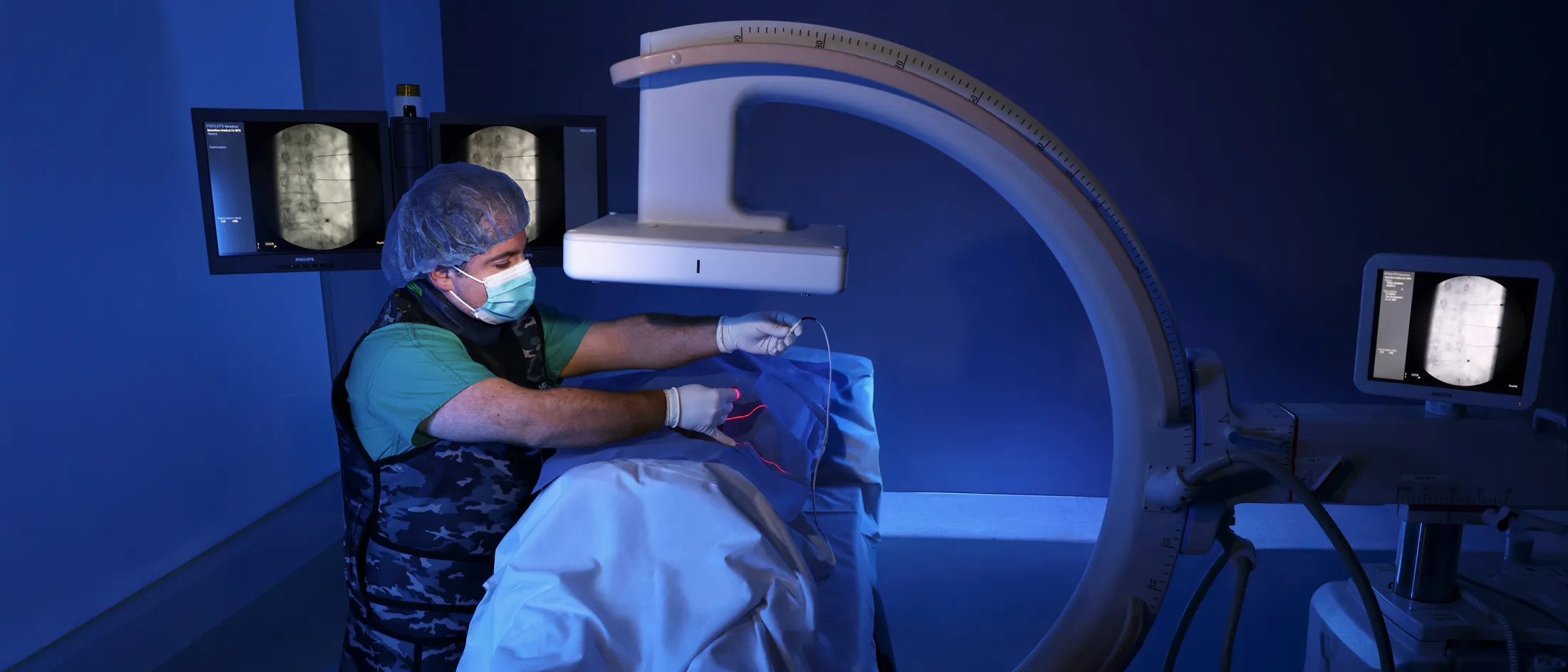 Physician in protective vest guiding a needle under C-arm fluoroscopy during a minimally invasive procedure.