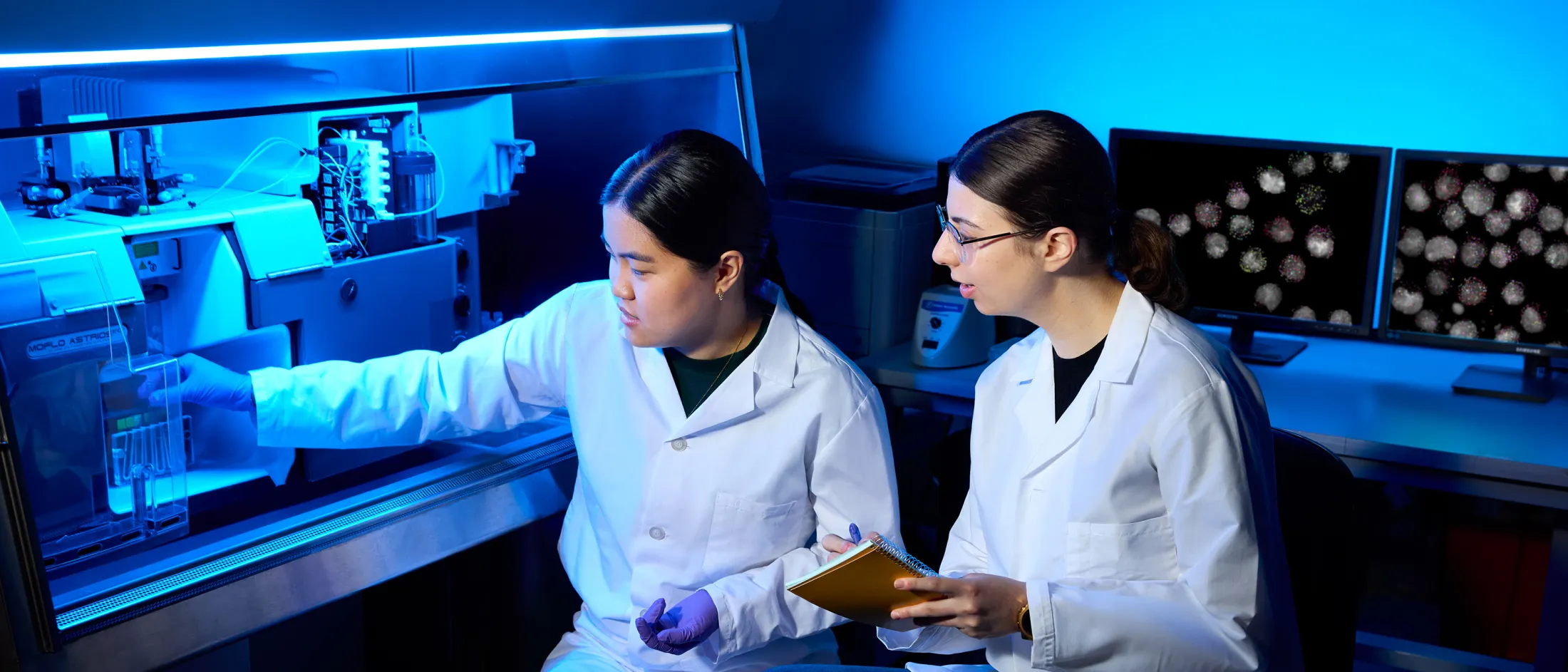 Two scientists in lab coats working with a large flow cytometer under blue lighting, with cell images displayed on monitors in the background.