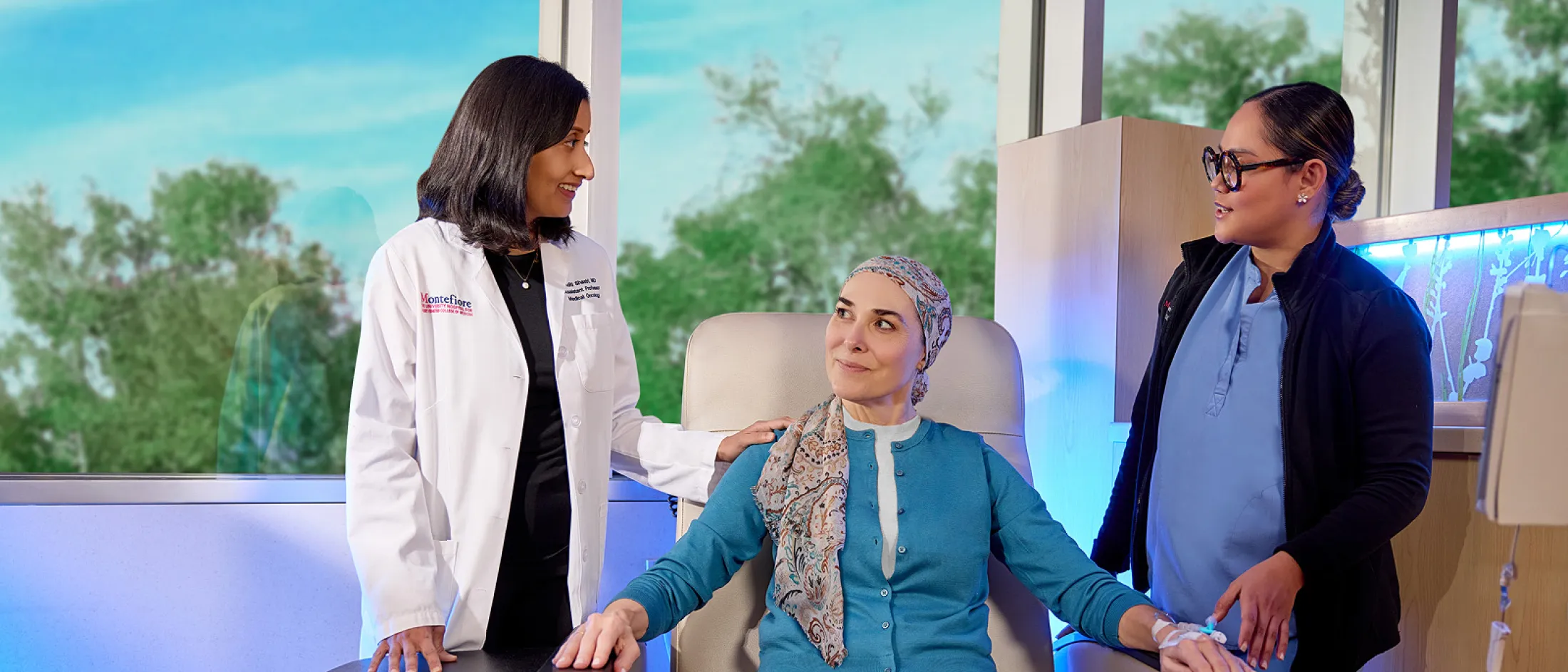 Female doctor and nurse comforting a woman receiving treatment, who is seated and wearing a headscarf in a clinical setting.