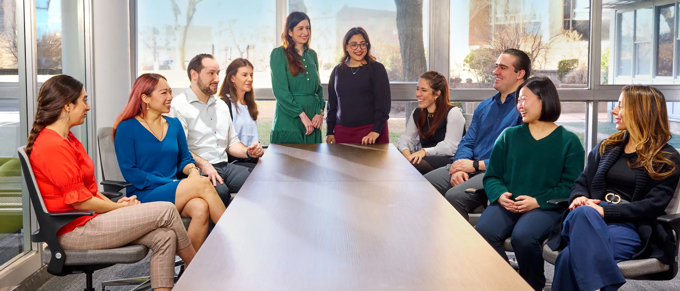 Large group of professionals seated and standing around a conference table, smiling in discussion near large windows
