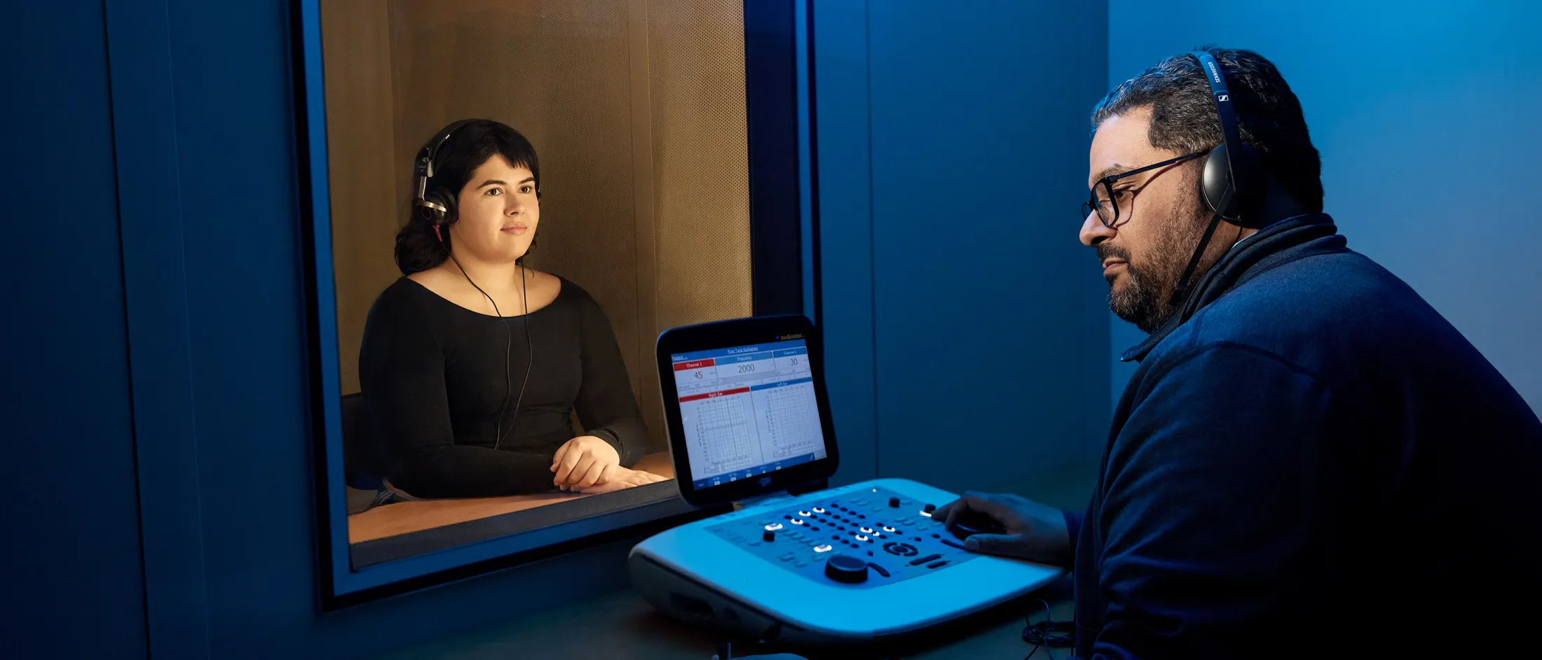 Woman in sound booth taking a hearing test while technician monitors data on a screen.