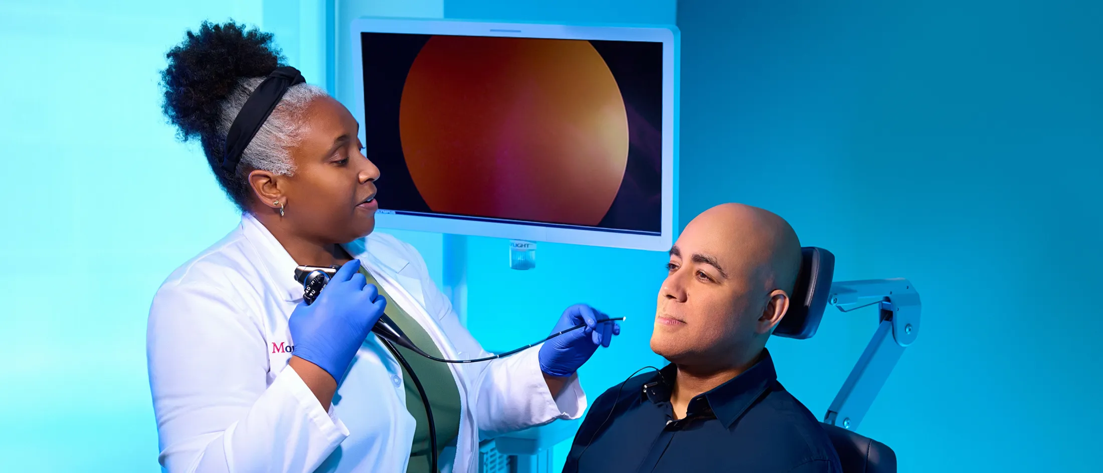 Doctor examining a patient’s throat using a flexible endoscope in a brightly lit medical exam room with a monitor display.