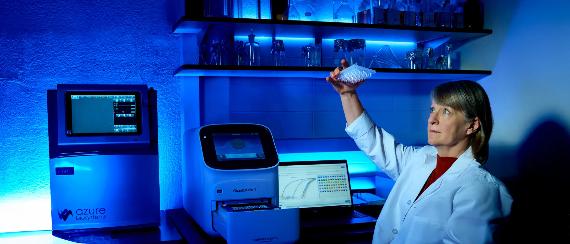 Female scientist in a lab coat inspecting a microplate in a laboratory with glowing blue ambient lighting and high-tech equipment.