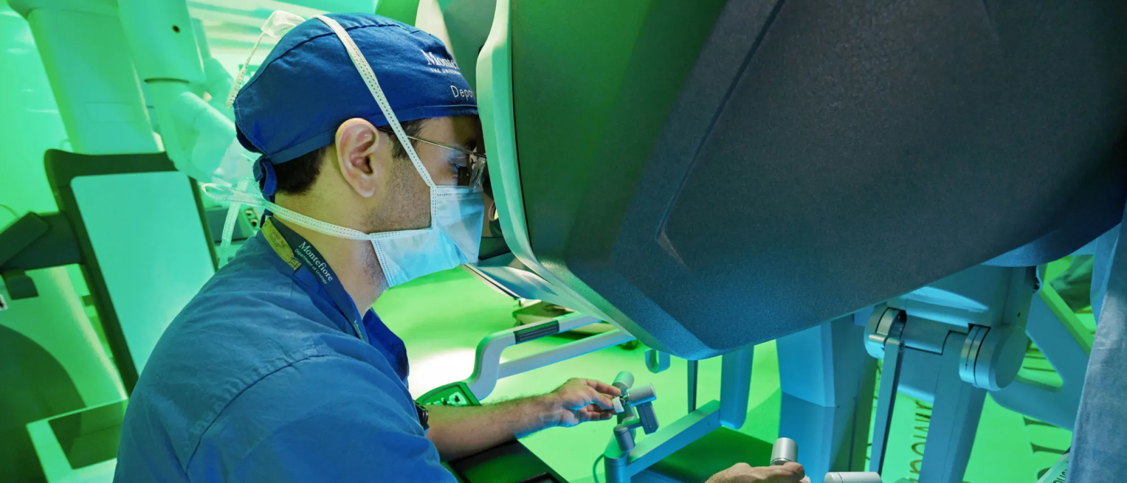 Surgeon operating robotic surgical system while looking through a console in a green-lit room.