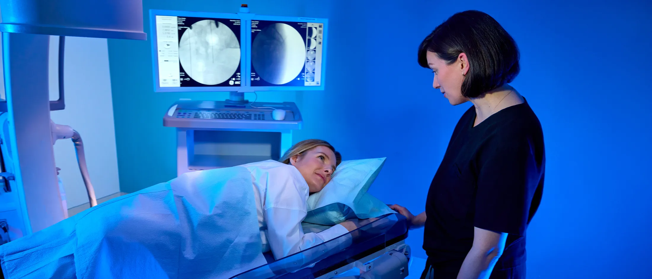 Healthcare provider talking with a patient lying on a medical imaging table with monitors in the background.