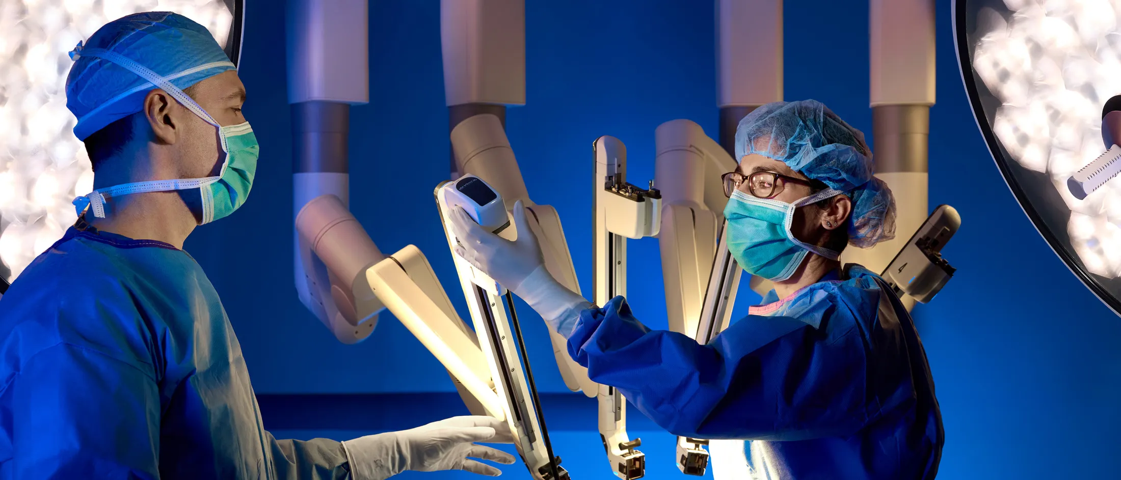 Two surgeons wearing scrubs and masks operate a robotic surgical system under bright lights in an operating room.