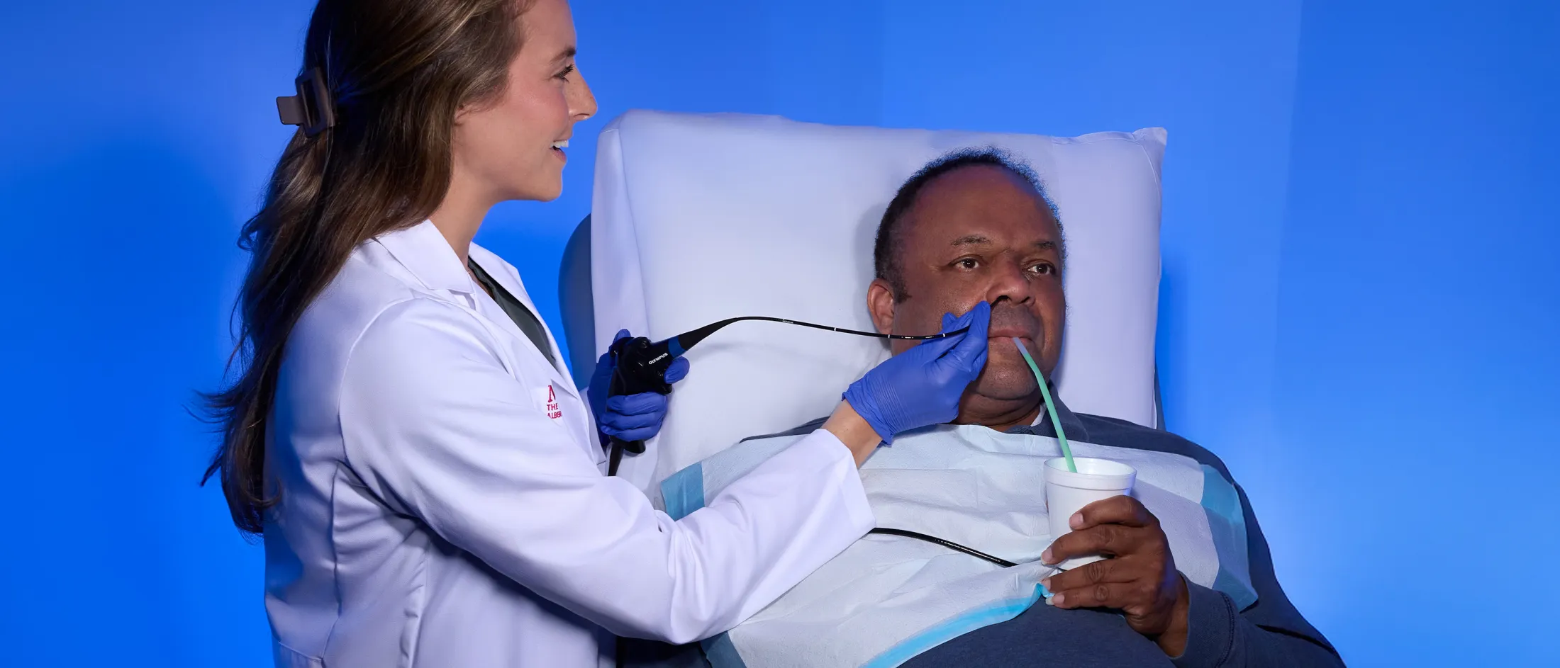 Female clinician performs a swallowing evaluation on a seated male patient using a flexible endoscope and a cup with a straw.