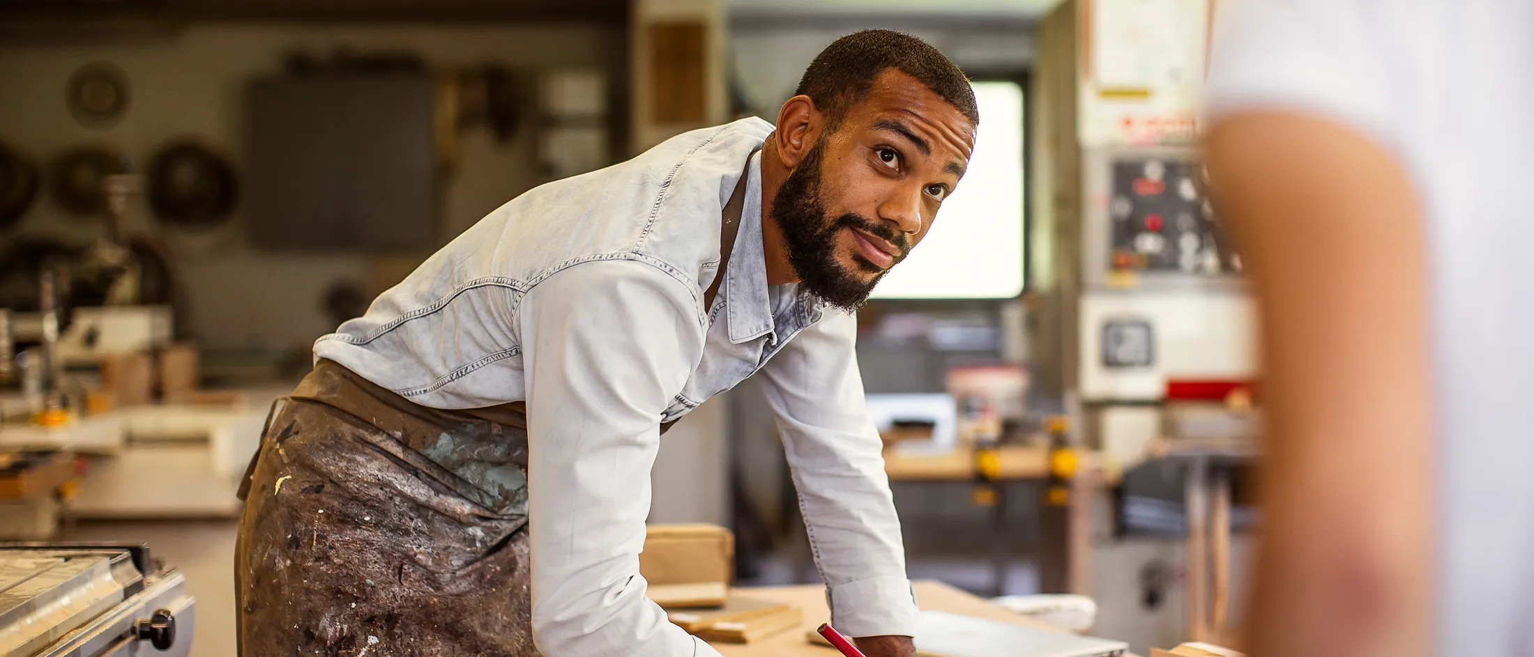 Man wearing a work apron leaning over a table and writing on paper in a woodshop, engaging with someone out of frame.