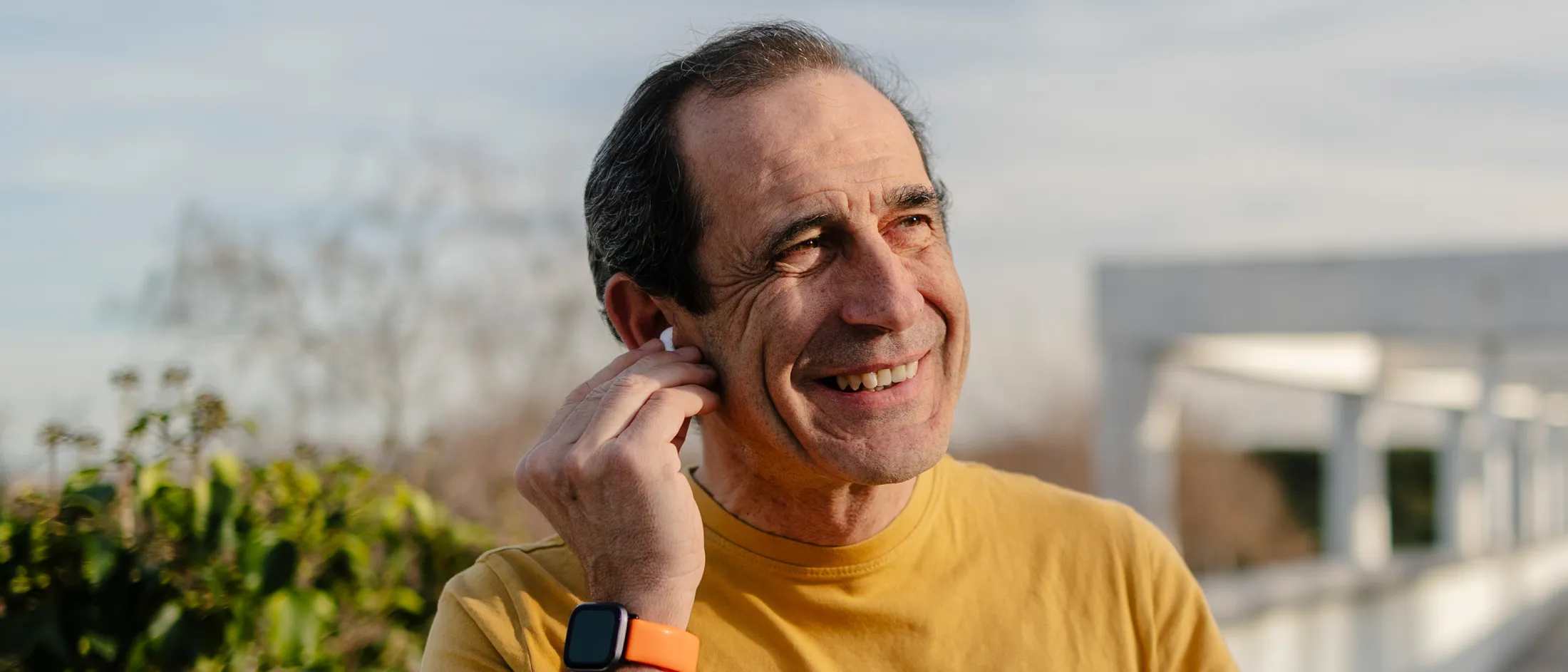 Older man smiles while adjusting his wireless earbud during a walk.