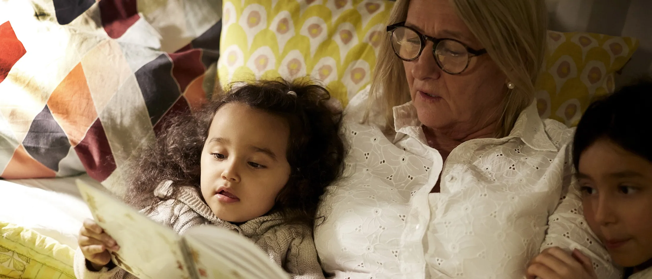 Older woman reading a picture book to two young children in a cozy bedroom setting.