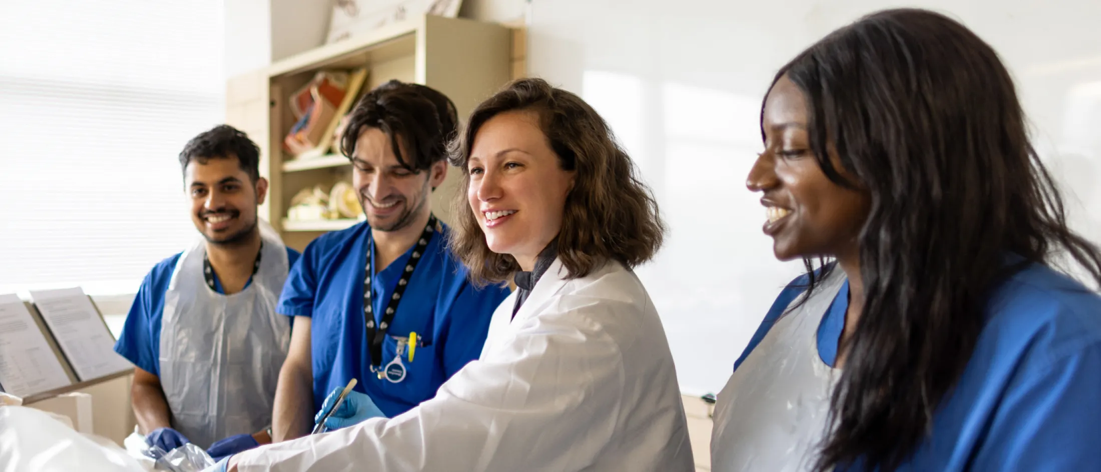 Medical instructor in a white coat demonstrating a procedure to three students in scrubs and aprons in a clinical classroom.