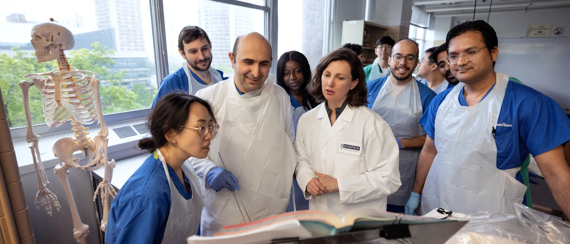 Group of medical students and instructors in lab attire gathered around a textbook and anatomical model in a classroom with large windows.