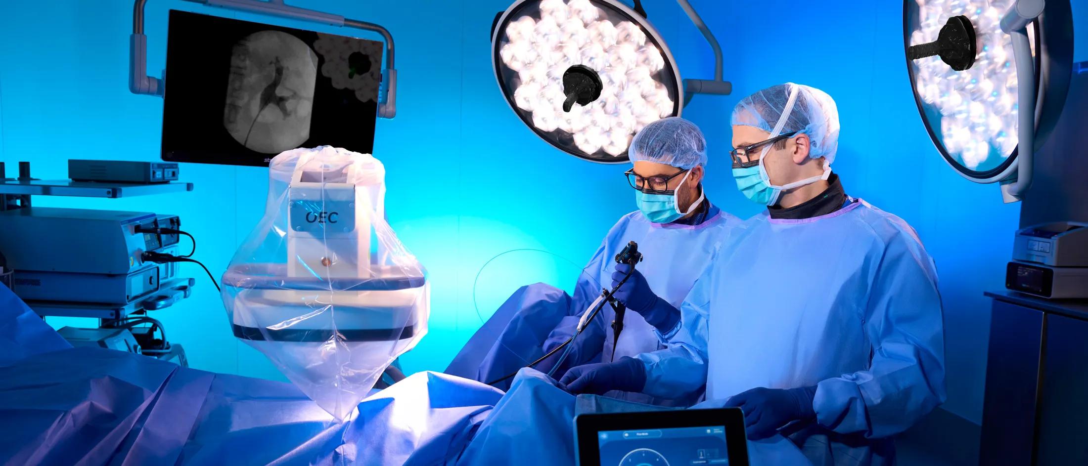 Two surgeons in an operating room perform a procedure under bright surgical lights with advanced imaging equipment.