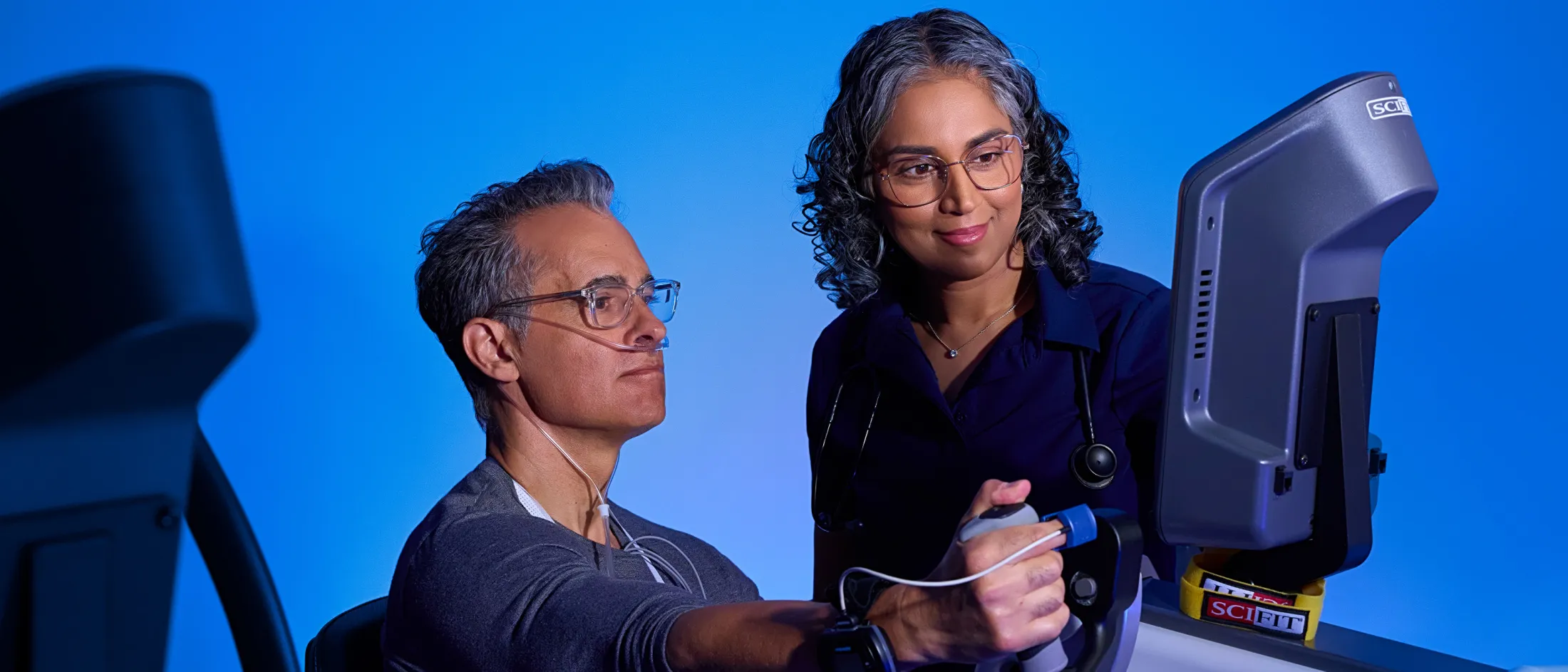 Woman supervising a man with a nasal cannula using an arm exercise machine during cardiopulmonary rehab.