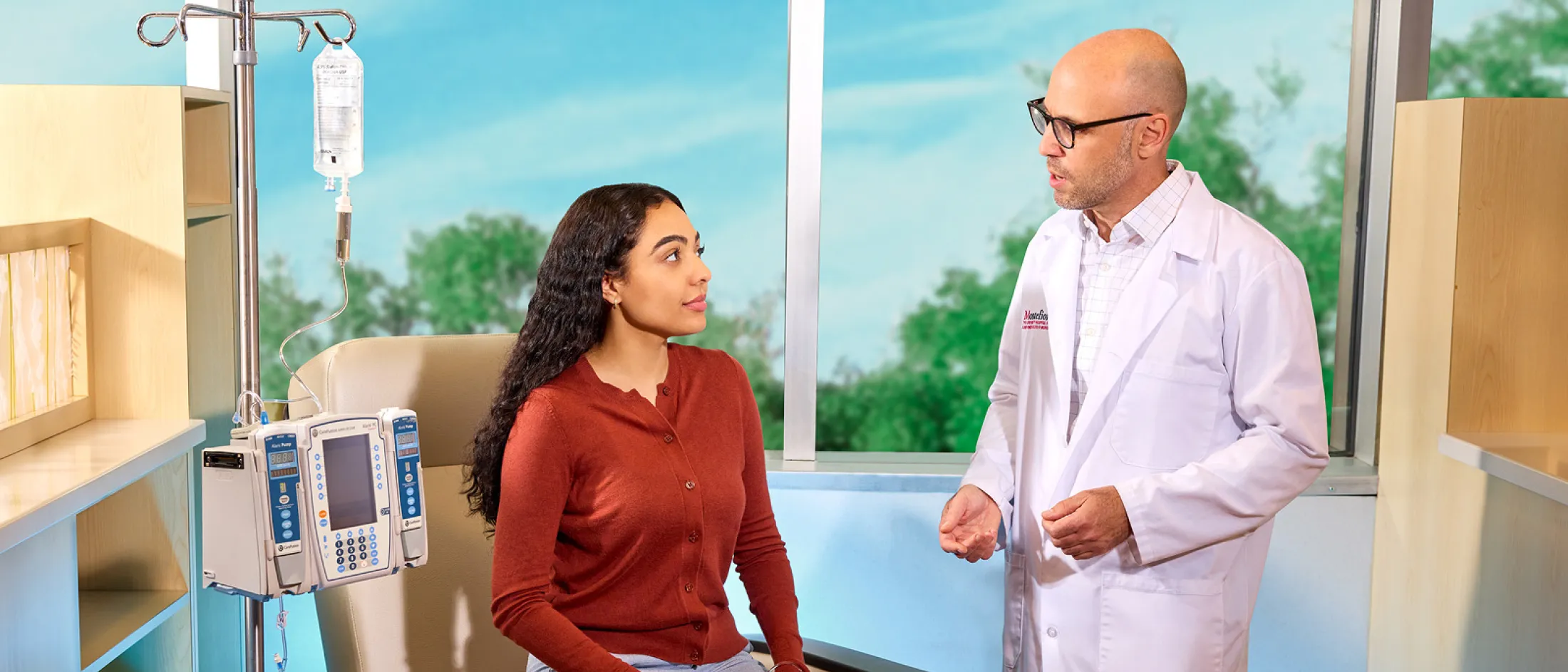 A patient receives IV treatment in a modern clinical setting while speaking with a doctor wearing a Montefiore Einstein lab coat.